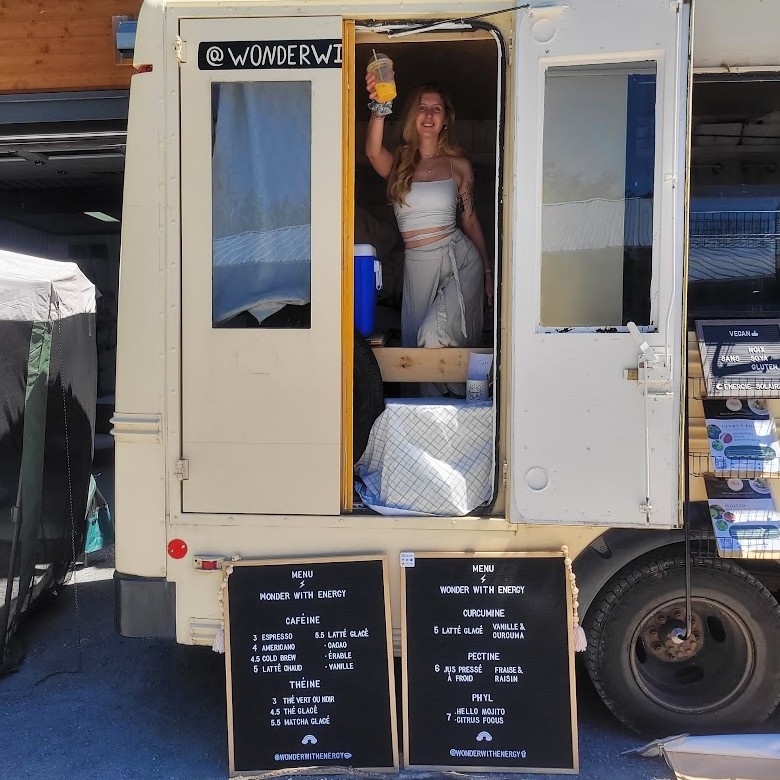 Woman smiling and holding up an orange drink from inside the Wonder with Energy bus, with menu boards visible.